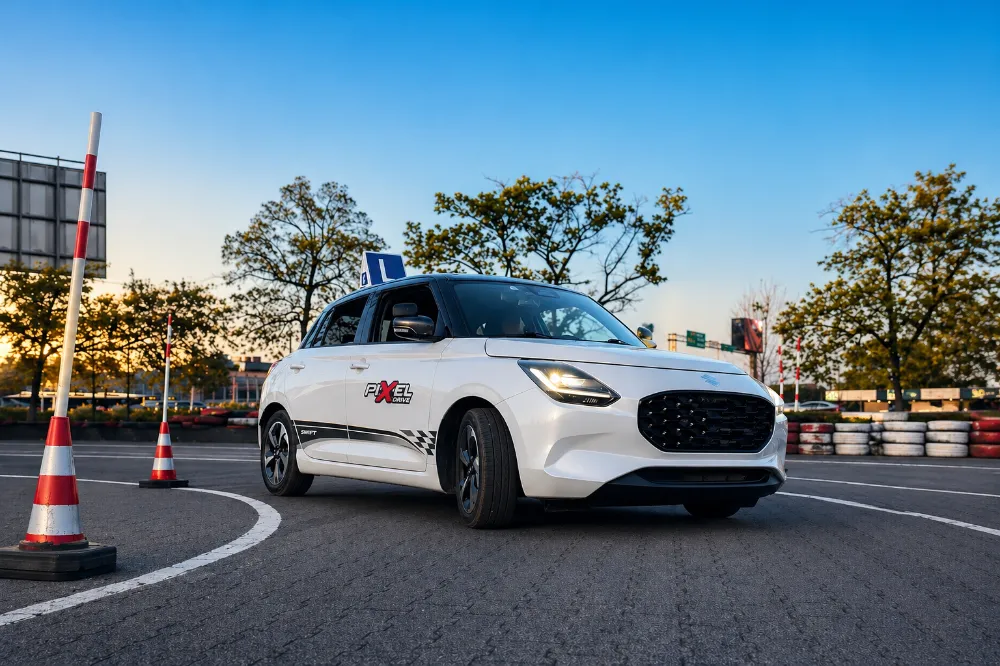 Biały Suzuki Swift szkoły jazdy PIXEL Drive z tablicą L między pachołkami na placu manewrowym o zachodzie słońca